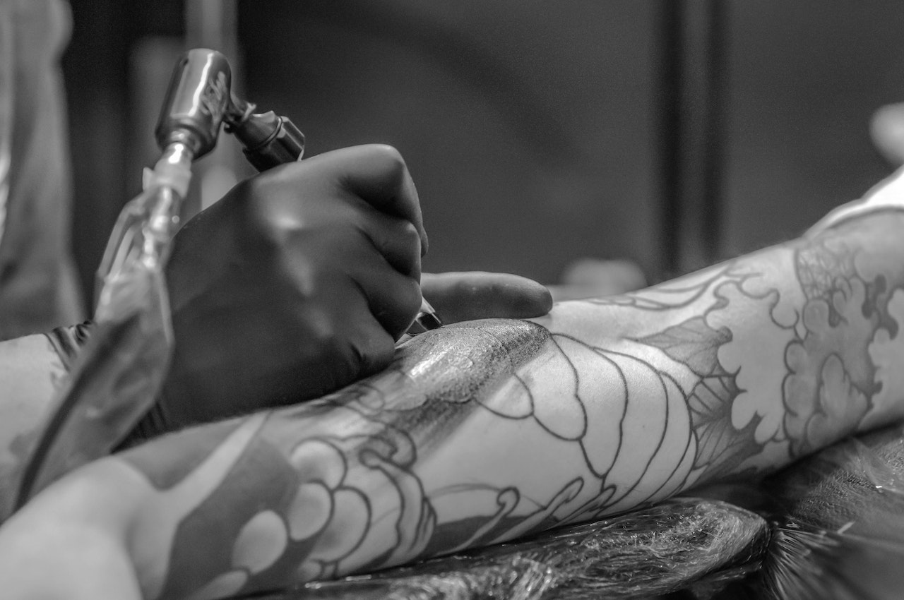 Na obrazku widoczny jest tatuażysta w pełnym skupienia, pracujący nad tatuażem złożonym z drobnych kropek na ciele swojego klienta. Jego precyzyjne ruchy igły tworzą wyjątkowy wzór, który nabiera życia pod jego biegłymi rękoma. Klient spokojnie leży, oddając się procesowi tworzenia tego minimalistycznego i oryginalnego tatuażu