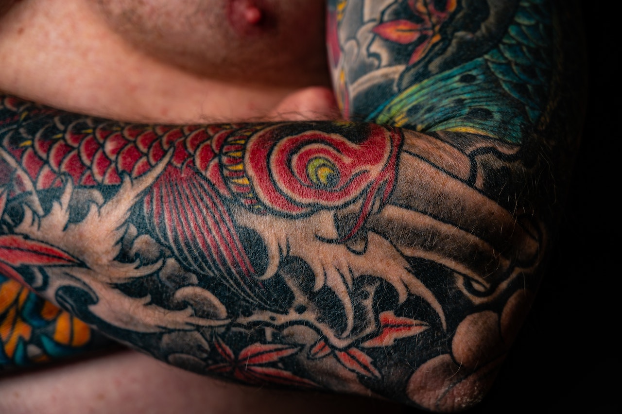 Na obrazku ukazuje się mężczyzna, którego skóra zdobi imponujący tatuaż w stylu japońskim. Wzór ten wpleciony jest w bogatą tradycję i symbolikę, tworząc na jego ciele piękną opowieść. Złożone detale i kolorowe elementy odzwierciedlają głębokie znaczenie tego tatuażu, podkreślając indywidualność i artystyczny duch noszącego go mężczyzny