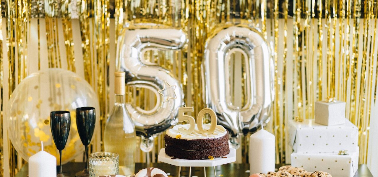 Na tym obrazku widoczna jest wspaniale przygotowana impreza z okazji 50. urodzin. Stół jest zastawiony pysznymi potrawami i tortem, ozdobami w kształcie liczby 50, a na środku stoi balon z liczbą "50". Goście cieszą się, bawią i składają życzenia jubilatowi, tworząc radosną i uroczystą atmosferę