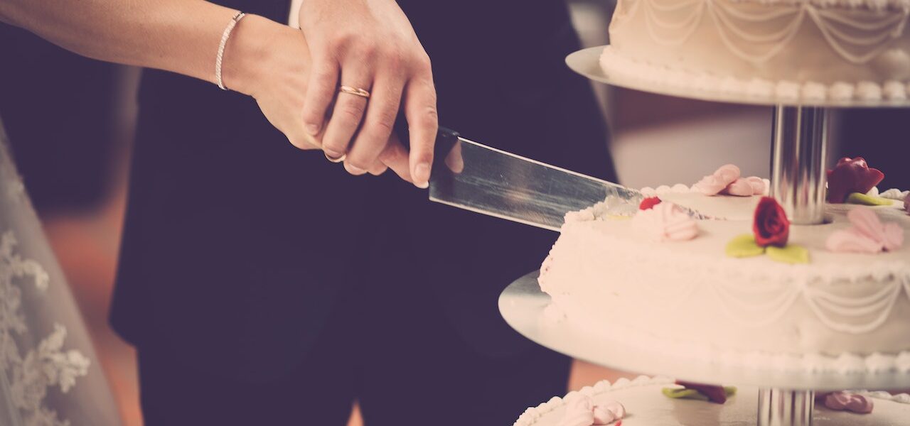 Na tym uroczym obrazku widoczna jest młoda para, która wspólnie kroi piękny tort weselny. Trzymają ręce na nożu i uśmiechają się do siebie, podczas gdy goście patrzą z zachwytem. To wzruszający moment, który symbolizuje ich wspólną podróż życiową i radość z tego wyjątkowego dnia