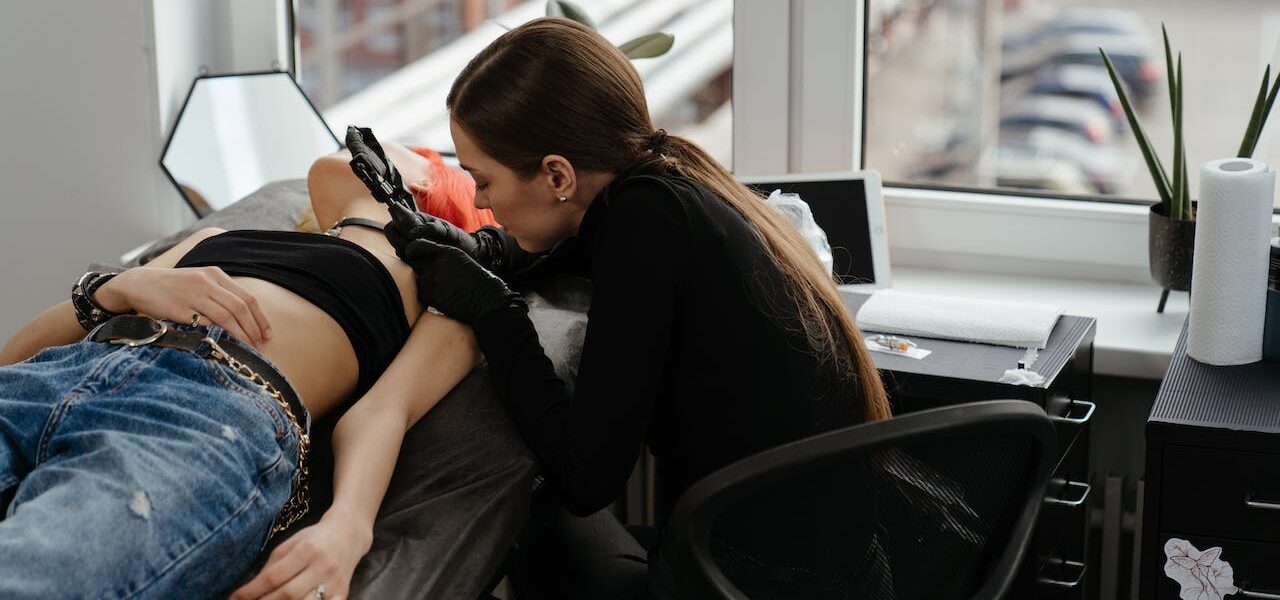Na obrazku, widzimy tatuażystkę, która precyzyjnie wykonywa realistyczny tatuaż na ramieniu swojej klientki. Jej zręczne ruchy i dbałość o szczegóły tworzą wrażenie trójwymiarowego dzieła sztuki